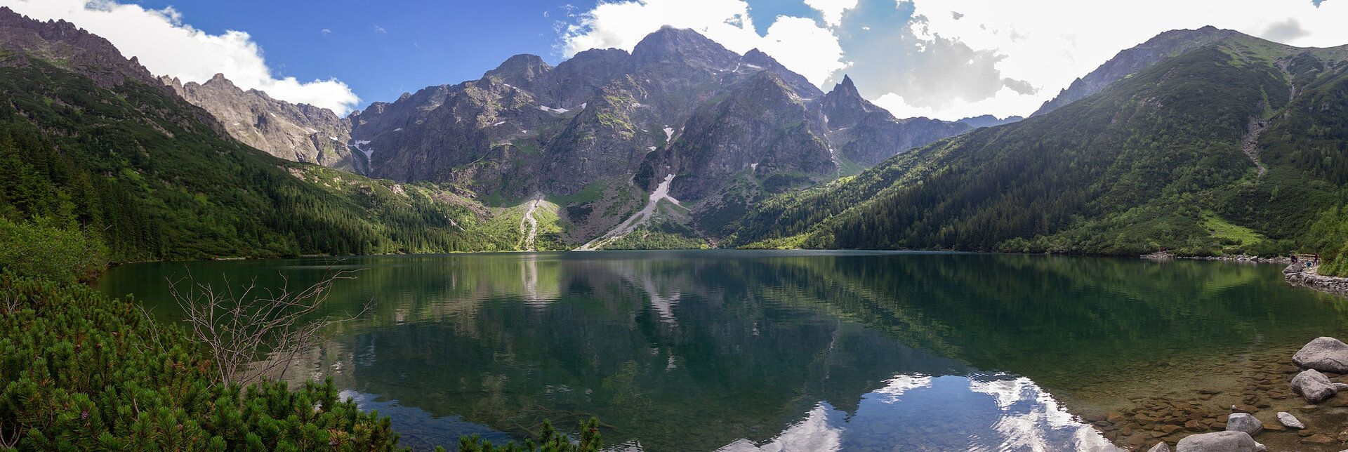 Fotografia jeziora otoczonego wysokimi, stromymi, skalistymi szczytami górskimi. W dolnych partiach pokryte drzewami, w górnych brak roślinności.