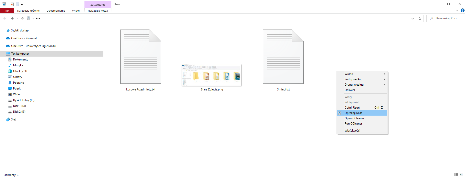Zrzut ekranu Eksploratora plików z otwartym folderem koszem. W prawym górnym rogu znajduje się nazwa bieżącego otwartego folderu "Moje prace", poniżej znajdują się zakładki "Pliki", "Narzędzia główne", "Udostępnianie", "Widok" oraz dwie zakładki zarządzania "Narzędzia Kosza", "Narzędzia obrazów". W drzewie folderów po lewej stronie znajdują się "Szybki dostęp",  "OneDrive - Personal", "OneDrive - Uniwersytet Jagielloński" oraz "Ten komputer" - "Dokumenty", "Muzyka", "Obiekty 3D", "Obrazy", "Pulpit", "Wideo", "Dysk lokalny (C:)", "Disk 1 (D:), "Disk 2 (E:)", "Sieć". W koszu znajdują się pliki "Losowe Przedmioty.txt", "Śmieci.txt" oraz "Stare Zdjęcie.jpg". W wyświetlonym menu kontekstowym dostępne są opcje "Widok", "Sortuj według", "Grupuj według", "Odśwież", "Wklej", "Wklej skrót", "Cofnij Usuń", "Opróżnij Kosz" (podświetlony), "Open CCleaner", "Run CCleaner",  "Właściowści".