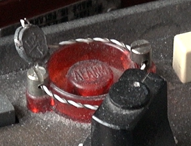 Zdjęcie przedstawia przycisk alarmowy radiostacji. Jest to duży okrągły czerwony przycisk z napisem Alarm. Jest on zabezpieczony przed przypadkowym wciśnięciem za pomocą druta owiniętego wokół przycisku.