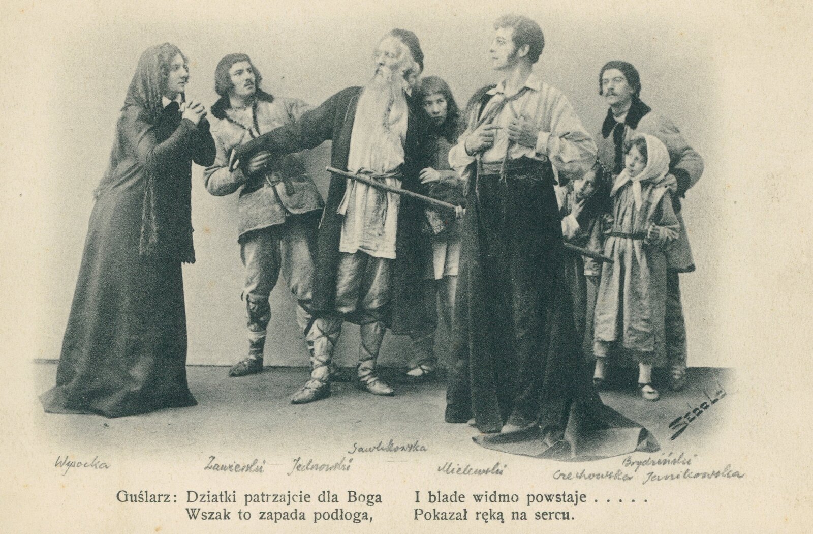 Kliknij, aby powiększyć. Zdjęcie przedstawia scenę z „Dziadów cz. II” Adama Mickiewicza – pojawienie się ostatniego ducha – Upiora. Kilku ludzi z pospólstwa przygląda się kobiecie i mężczyźnie. Jednym z nich jest Guślarz, postać, która przewodniczyła obrzędowi dziadów. Człowiek ten wyróżnia się starszym, niż pozostali, wiekiem i długą brodą. Dwoje wpatrujących się w siebie ludzi – kobieta i mężczyzna – nie widzi poza sobą świata. Ona składa ręce jak do modlitwy, on jest duchem przybyłym na dziady, by spotkać się z dawną ukochaną. Trzyma ręce na sercu. Na dole zdjęcia widoczny jest fragment z dramatu: Guślarz: Dziatki patrzajcie na Boga/ Wszak to zapada podłoga,/ I blade widmo powstaje…/ Pokazał ręką na serce. Powyżej tego tekstu są wypisane nazwiska aktorów grających w spektaklu Dziadów poszczególne role.