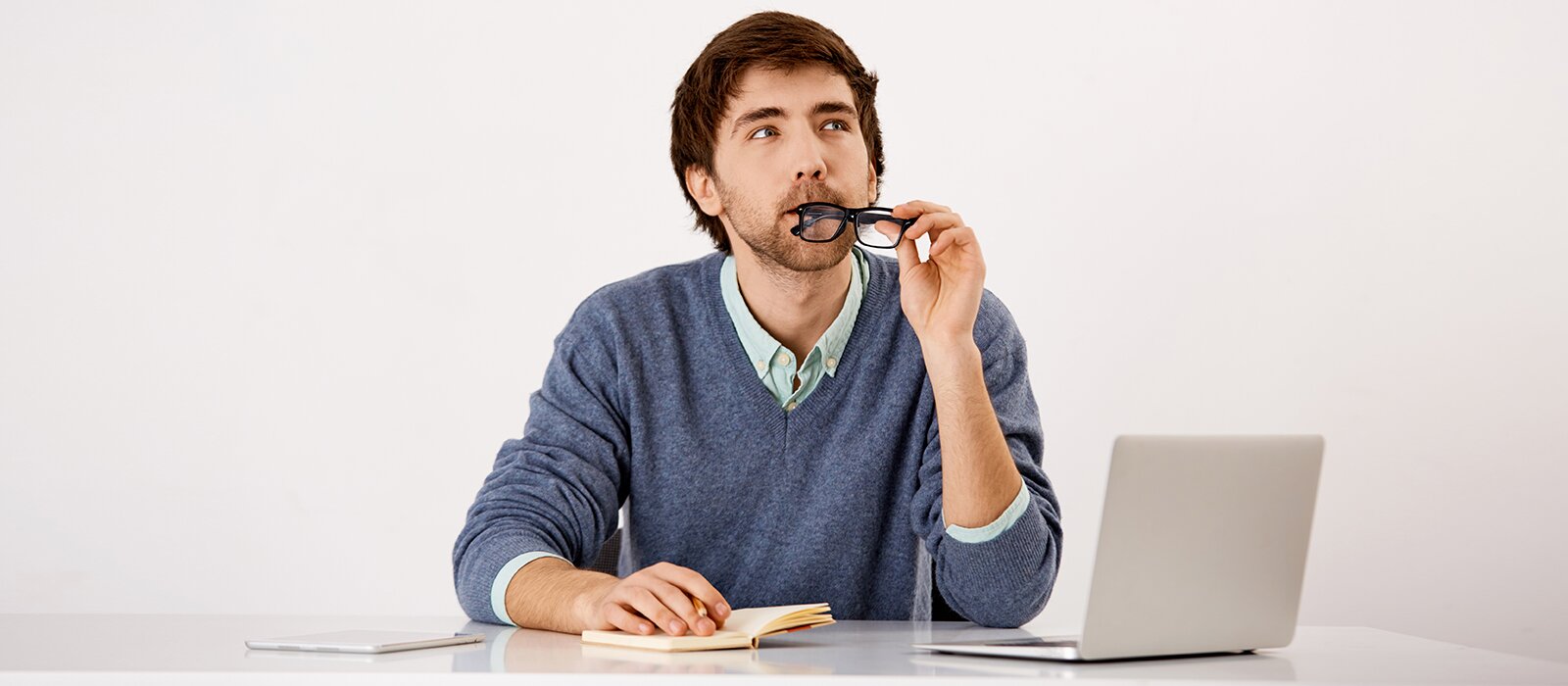 Zdjęcie przedstawia młodego mężczyznę siedzącego za blatem. Mężczyzna ma krótkie, ciemne włosy, jest ubrany w jasną koszulę i niebieski sweter. Głowę ma zwróconą lekko w górę w prawo. W lewej dłoni trzyma okulary korekcyjne, które  przygryza ustami. Prawą dłoń ma położoną na notatniku leżącym na blacie. Przed nim jest widoczny otwarty laptop oraz otwarty notatnik.