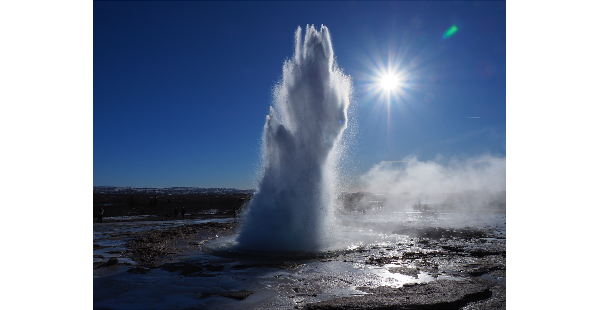 Zdjęcie przedstawia wybuch gejzera pośród błota na dużą wysokość. Niebo bezchmurne, błękitne, z widocznym Słońcem. Wybuch widoczny jako szara plama o owalnym kształcie.