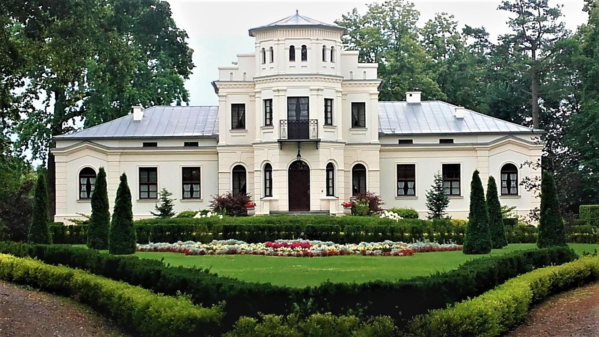 Zdjęcie przedstawia biały, parterowy dworek z szarym dachem. W jego centralnej części znajduje się dwupiętrowa wieża. Okna w budynku rozmieszczone są symetrycznie. Za dworkiem rosną wysokie, zielone drzewa. Przed budynkiem znajduje się zadbany ogród, w którym rosną przycięte choinki i krzewy, zielona trawa, a także białe i czerwone kwiaty. Niebo jest jasne. Zdjęcie zostało wykonane w świetle dziennym.