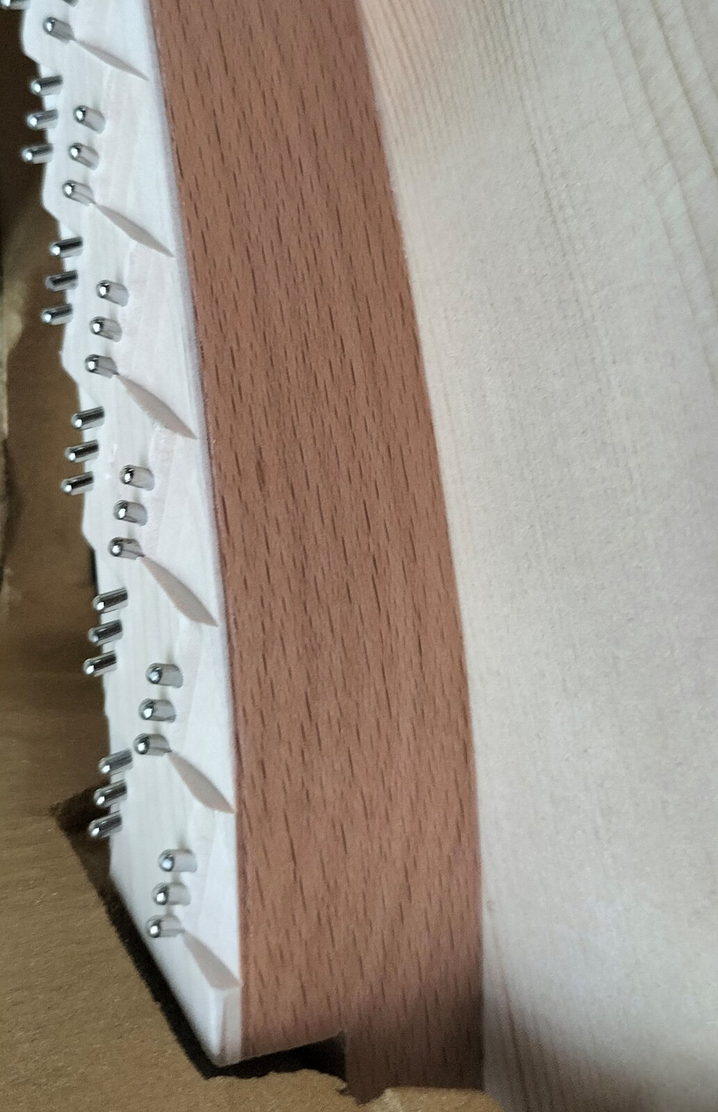 Zdjęcie przedstawia zbliżenie na próg wiolinowy wykonany z drewna bukowego. Po lewej stronie widoczny jest biały pas ze srebrnymi kołkami stroikowymi, na prawo którego znajduje się delikatnie łukowaty pas z brązowej sklejki. Po prawej widoczny jest fragment progu z jasnobeżowego drewna bukowego z widocznymi cienkimi paskami wzdłuż obrazującymi strukturę drewna.