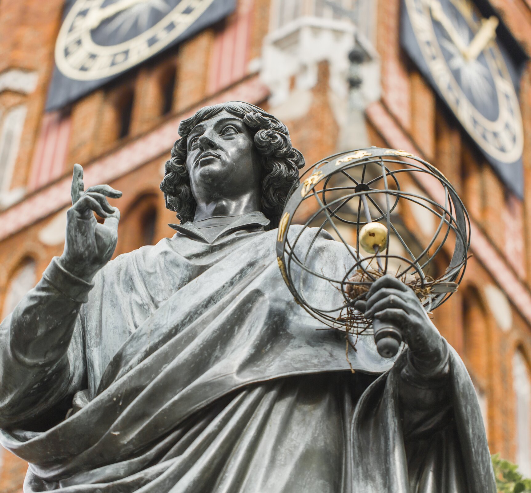 Ilustracja przedstawia fragment pomnika Mikołaja Kopernika znajdującego się w Toruniu. Monument przedstawia Kopernika ubranego w togę uczelnianą, który w lewej ręce trzyma sferę armilarną, a palcem prawej ręki wskazuje niebo. W tle budynek z dużymi dwoma zegarami.