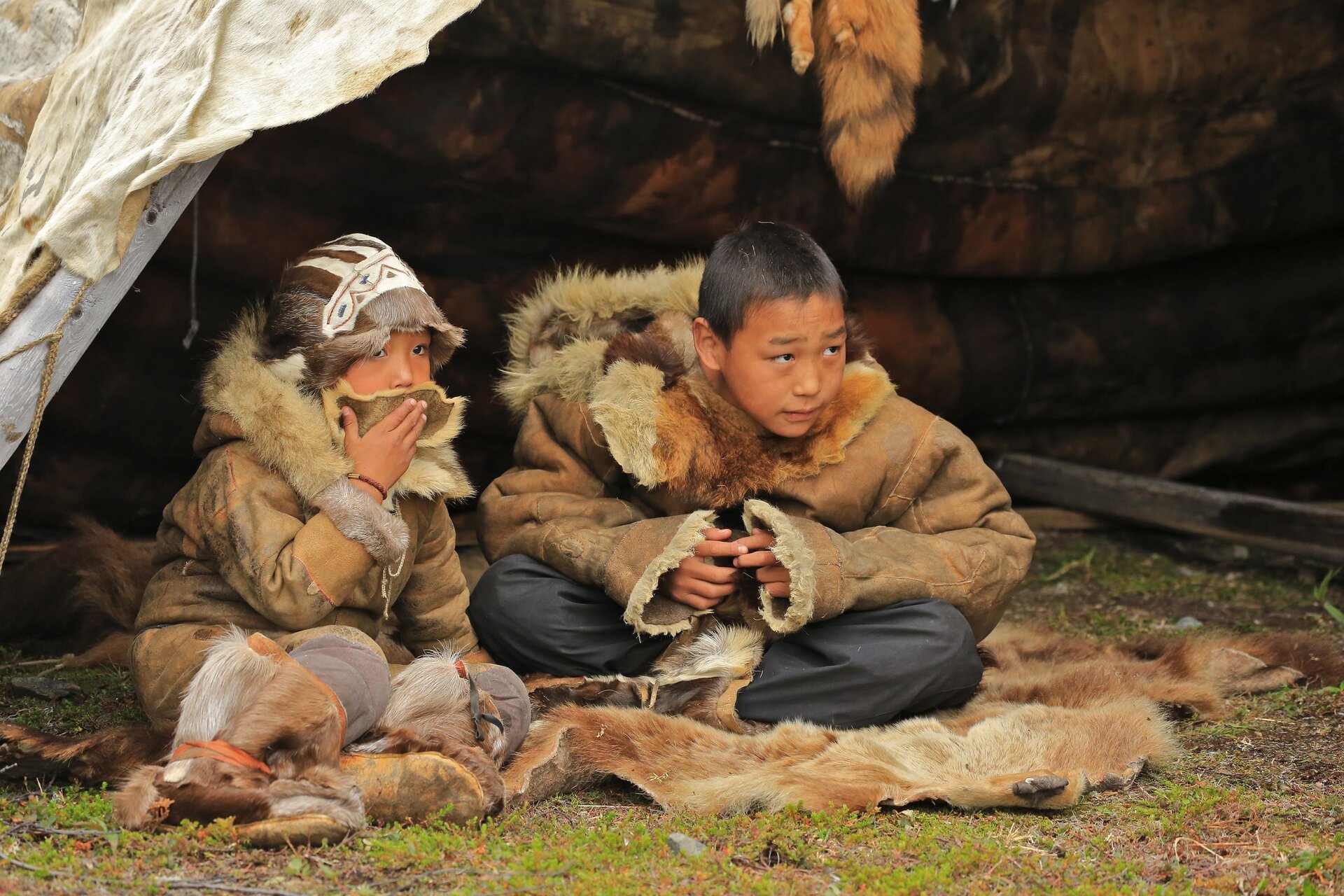 Zdjęcie przedstawia dwoje dzieci ubranych w kożuchy wywrócone skórą do spodu. Siedzą na rozłożonej na ziemi skórze. 