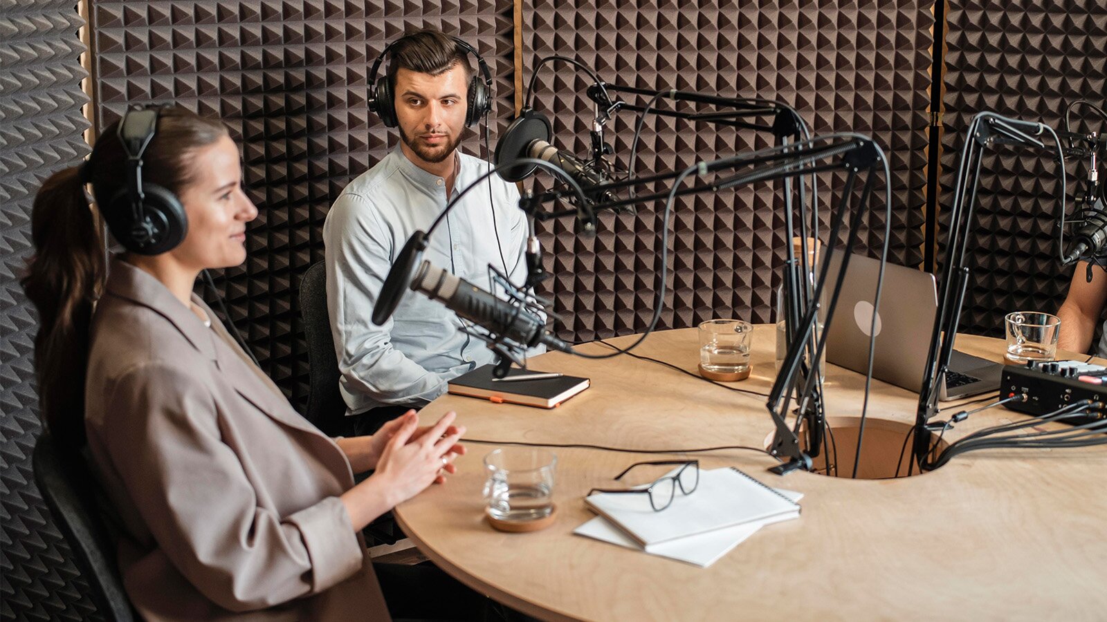 Zdjęcie przedstawia kobietę oraz siedzącego obok mężczyznę. Para siedzi w słuchawkach przy stole, nad którym wiszą mikrofony na statywach. Ściana za nimi jest wyłożona pianką wygłuszającą. 