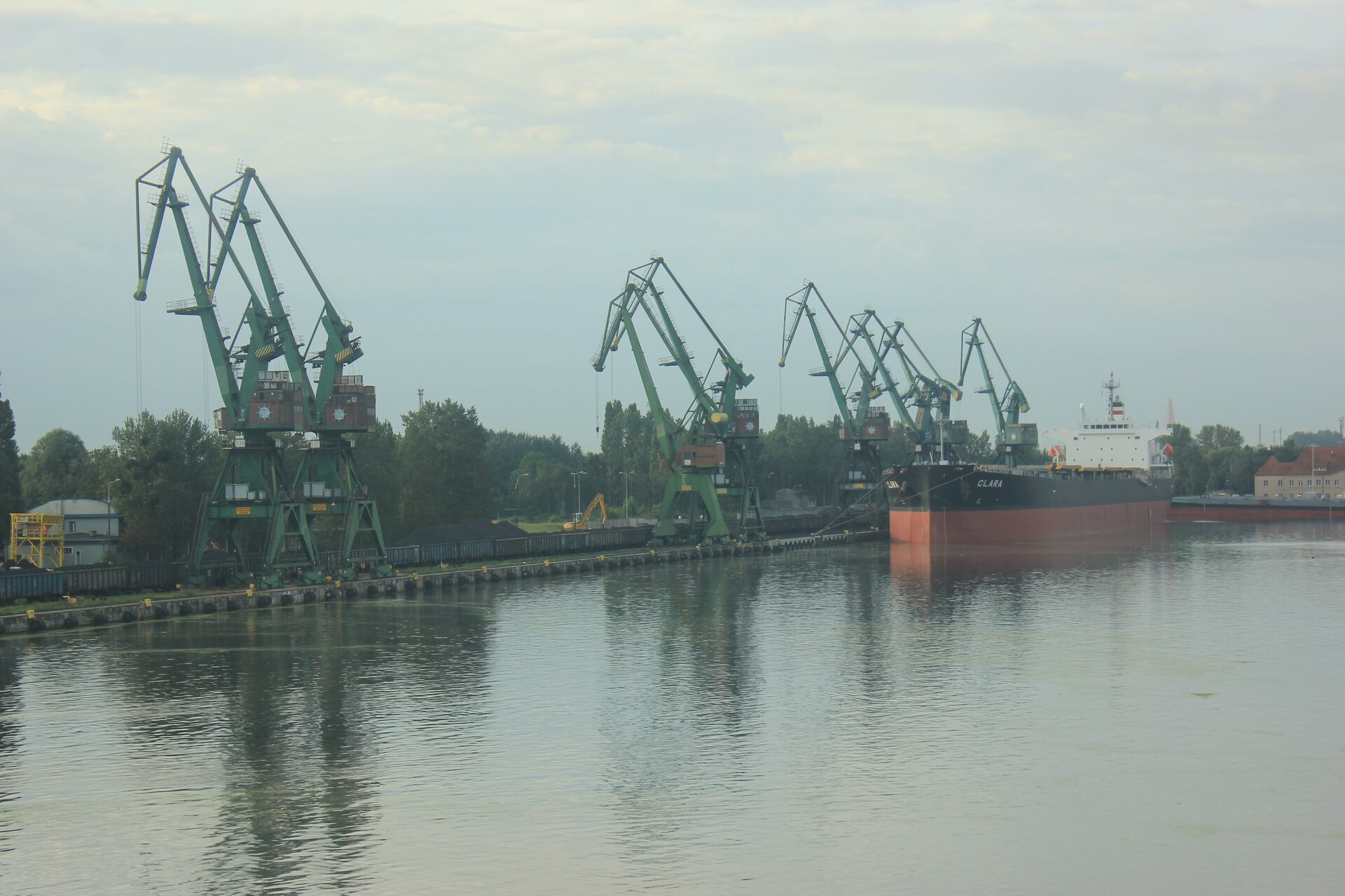 Fotografia kilku wysokich żurawi w porcie. Obok nich do brzegu zacumowany statek towarowy. Na pierwszym planie woda. Za żurawiami linia drzew.