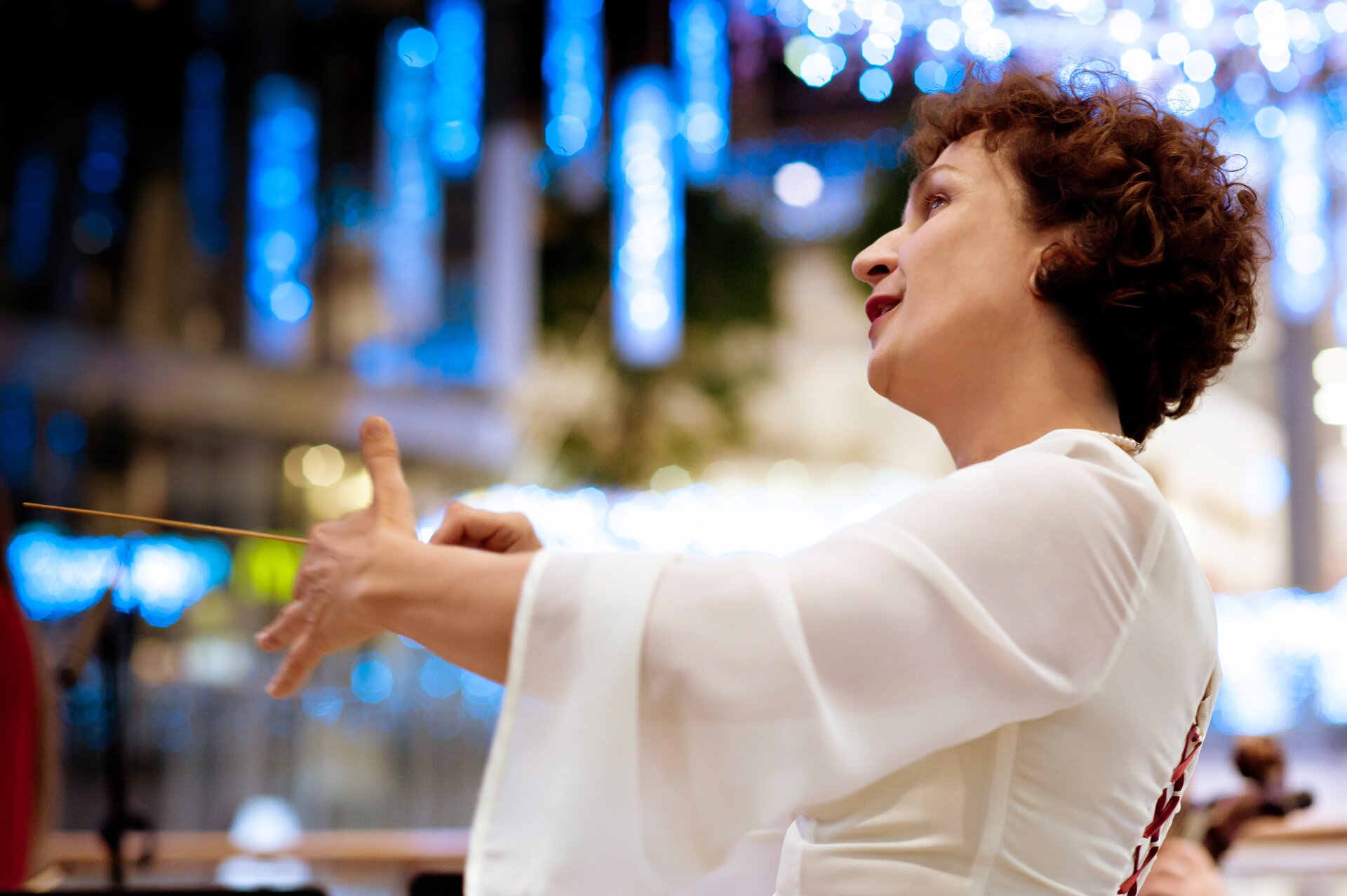 Zdjęcie przedstawia dyrygującą kobietę. W prawej dłoni trzyma batutę. Drugą ręką wykonuje gest. Głowę ma przechyloną na bok. Ma na sobie białą koszulę. W tle znajdują się niebieskie światła.