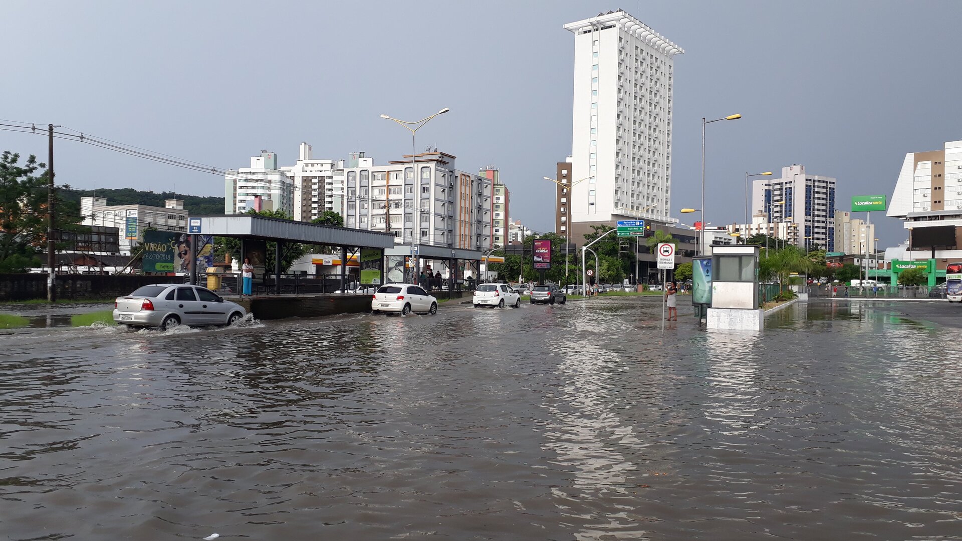 Zalana ulica w mieście. Poziom wody osiąga wysokość około dwudziestu centymetrów, co utrudnia ruch samochodów oraz pieszych. Na drugim planie kilkunastopiętrowe bloki mieszkalne.