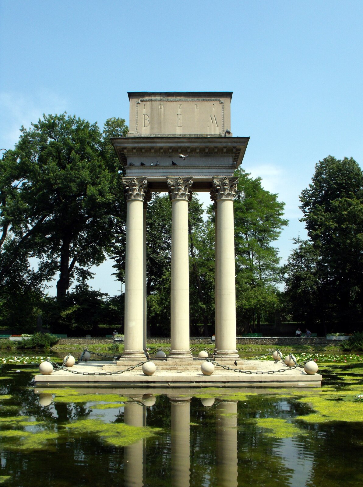 Zdjęcie przedstawia grób w postaci trzech wysokich kolumn z portykiem, na którym znajduje się napis: Józef Bem. Szczyty kolumn posiadają roślinne ornamenty. Kolumny otoczone są kamiennymi kulami połączonymi łańcuchami. Wokół konstrukcji jest staw. W tle rosną drzewa.