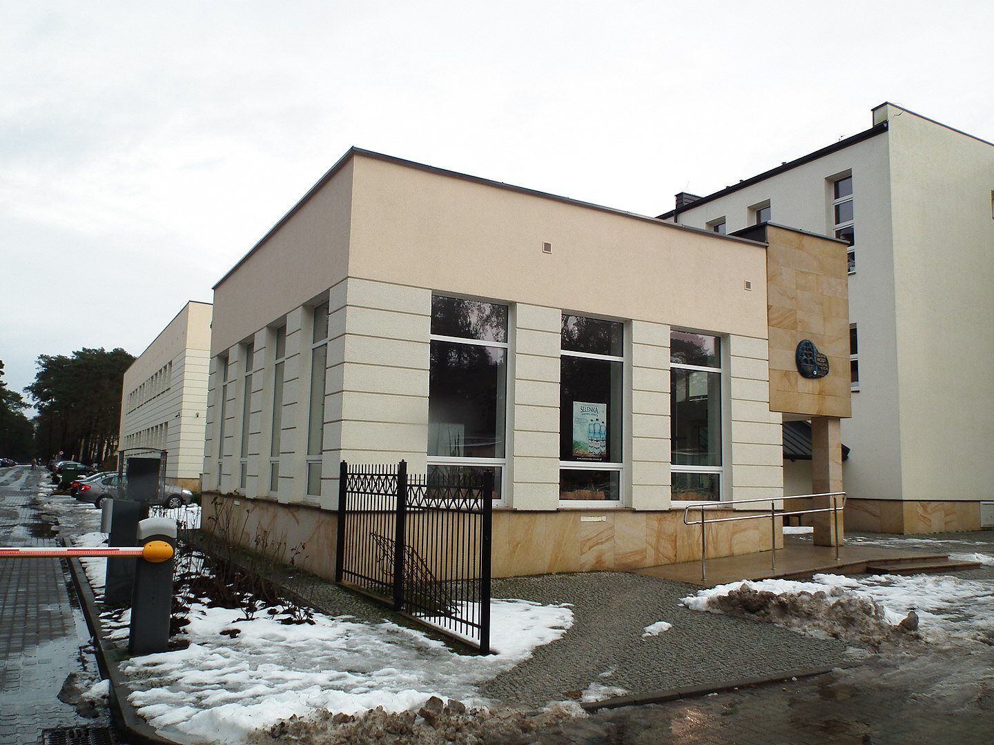 Zdjęcie przedstawia kompleks prostokątnych budynków. Budynek na pierwszym planie ma duże okna. Na chodniku oraz na drodze leży śnieg.