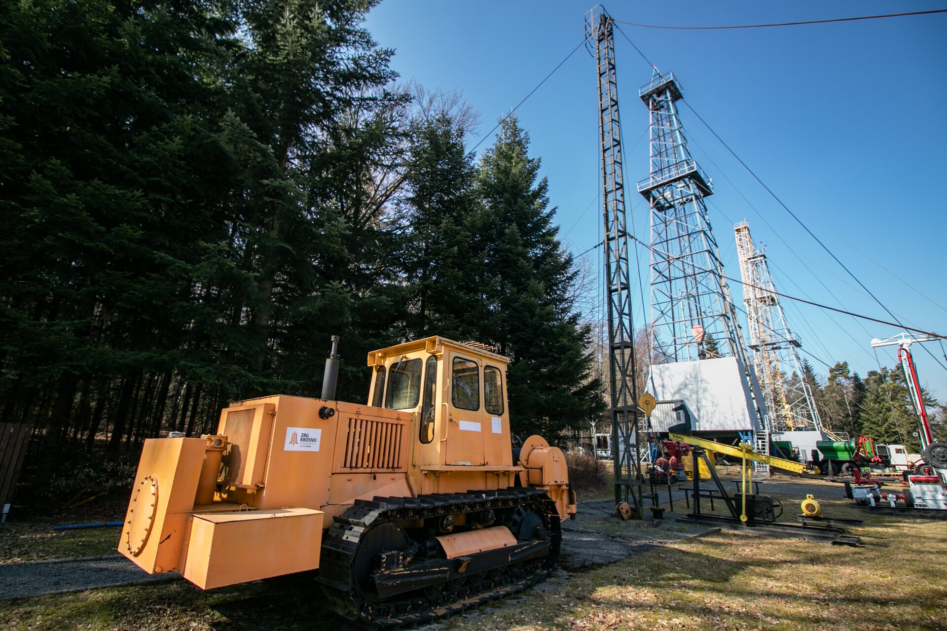 Na zdjęciu przedstawiona jest winda wyciągowa typu <math aria‑label="Bakiniec trzy">Bakiniec 3. Jest to pomarańczowy pojazd gąsienicowy przypominający połączenie traktora z walcem drogowym. W tylnej części zamiast walca posiada on bęben. Na bęben nawiniętą jest czarna lina. Winda z przodu ma dwa prostopadłościenne elementy. Na górze obudowy windy widać rurę wydechową. Za pojazdem znajduje się wysoki metalowy maszt. Jest to wiertnia. Do obu stron masztu przymocowane są po dwie liny odciągowe. Liny zamontowane są na szczycie i w połowie wysokości masztu. Obok masztu zainstalowany jest kiwon pompowy. W górnej części znajduje się żółte ramię wahacza. Z lewej strony ramienia znajduje się jego łeb, do którego przyłączona jest linka zwana żerdzią dławikową. Linka poprowadzona jest w linii prostej do głowicy odwiertu. Głowica odwiertu jest walcem wystającym z ziemi.  Od niej z lewej strony poprowadzona jest czarna rurka idąca do ziemi. Ramię wahacza umieszczone jest na czarnej ramie nośnej. Rama nośna jest w kształcie ostrosłupa. Obok ramy nośnej znajduje się prostokątny element z dwoma żółtymi ramionami. Jest to pociągacz. Pociągacze są przyłączone do wahacza. Do czarnego elementu będącego mechanizmem kiwona przytwierdzone jest koło zamachowe. Najbardziej na prawo znajduje się znajduje się żółty okrągły silnik. W oddali widać dwa duże słupy przypominające słupy sieci wysokiego napięcia, kilka pojazdów oraz drzewa.