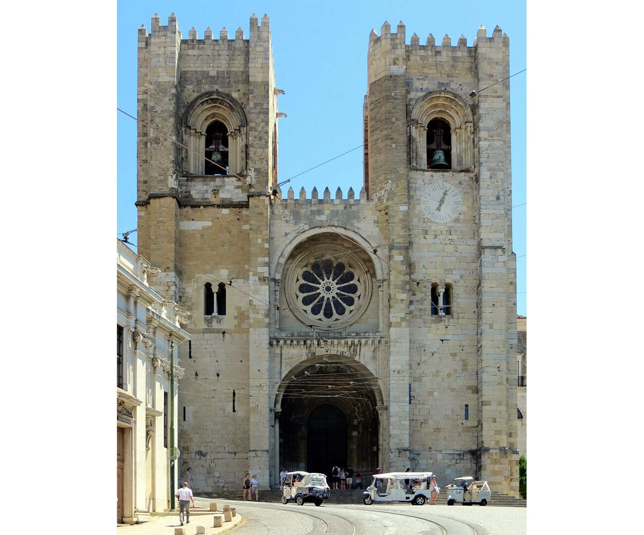 Fotografia barwna przedstawiająca Katedrę w Lizbonie w  Portugalii, w połowie XII wieku. Katedra zbudowana na kształt dwóch wieży. Ozdobione zewnętrznie okna z wyżłobionymi elementami. Po środku, pomiędzy wieżami wielkie okrągłe okno- witraż. Pod nim ogromna brama wejściowa.