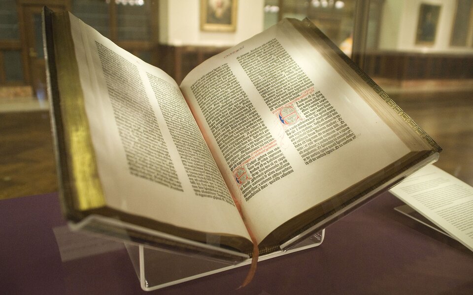 Kliknij, aby powiększyć Zdjęcie przedstawia otwartą biblię. Pośrodku jest cienki pasek, stanowiący zakładkę. Tekst napisany jest na każdej stronie w dwóch kolumnach. Grzbiet książki jest koloru złotego. Książka jest na plastikowym stojaku. Po prawej stronie jest karta z zapisanym tekstem.