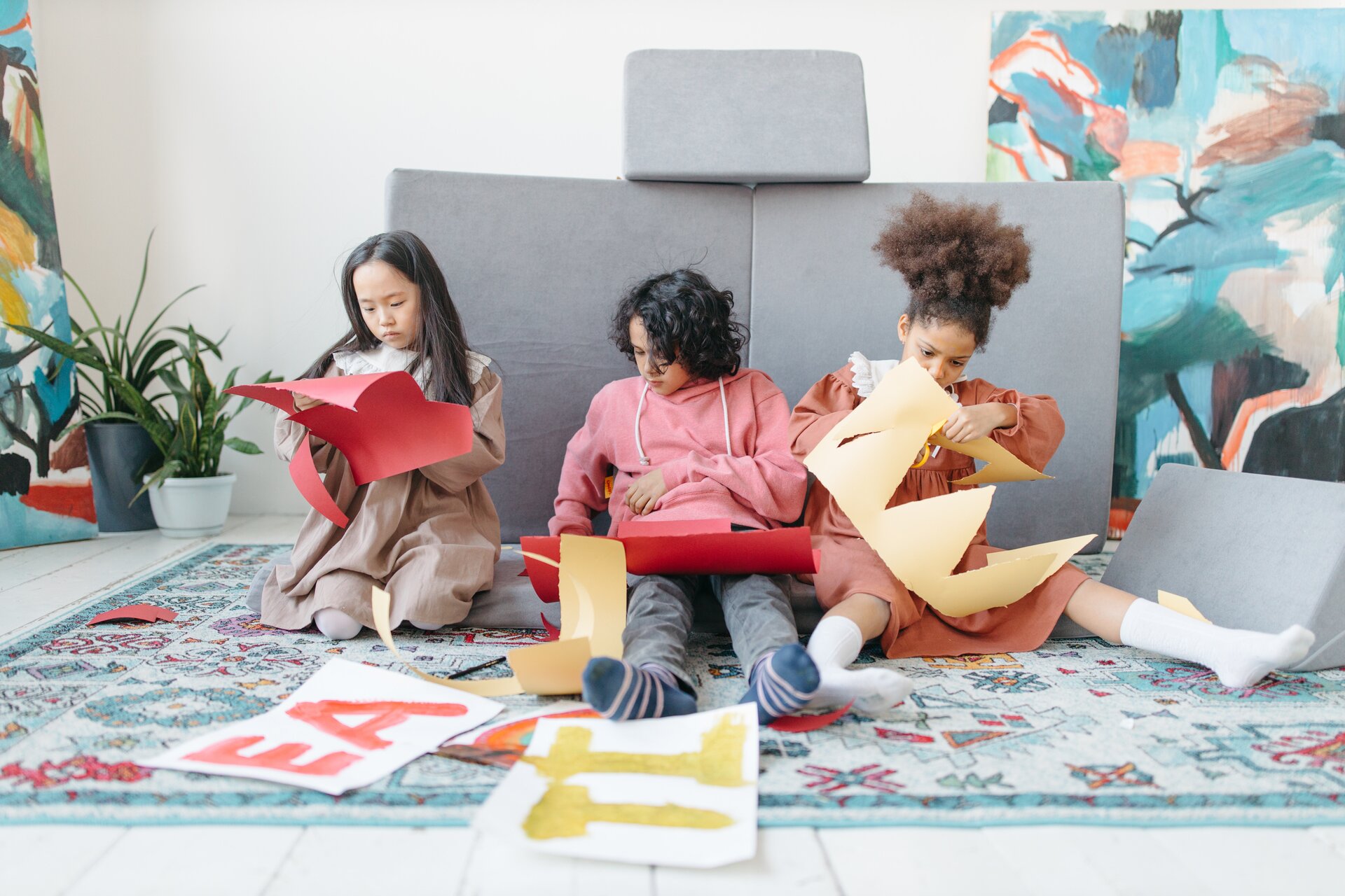 Zdjęcie przedstawia troje dzieci z różnych regionów świata. Siedzą na dywanie i wycinają litery z papieru. 