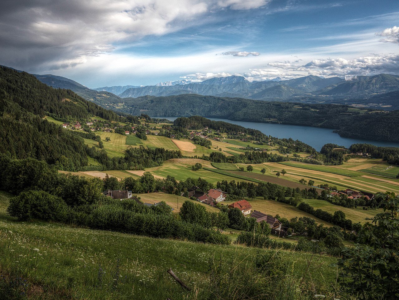 Ilustracja przedstawia krajobraz wsi w Austrii. W tle znajdują się góry, gdzieniegdzie pokryte śniegiem. Zbocza wzgórz na pierwszym planie pokryte są drzewami iglastymi. Na terenach równinnych znajdują się pola uprawne, między nimi domy. 