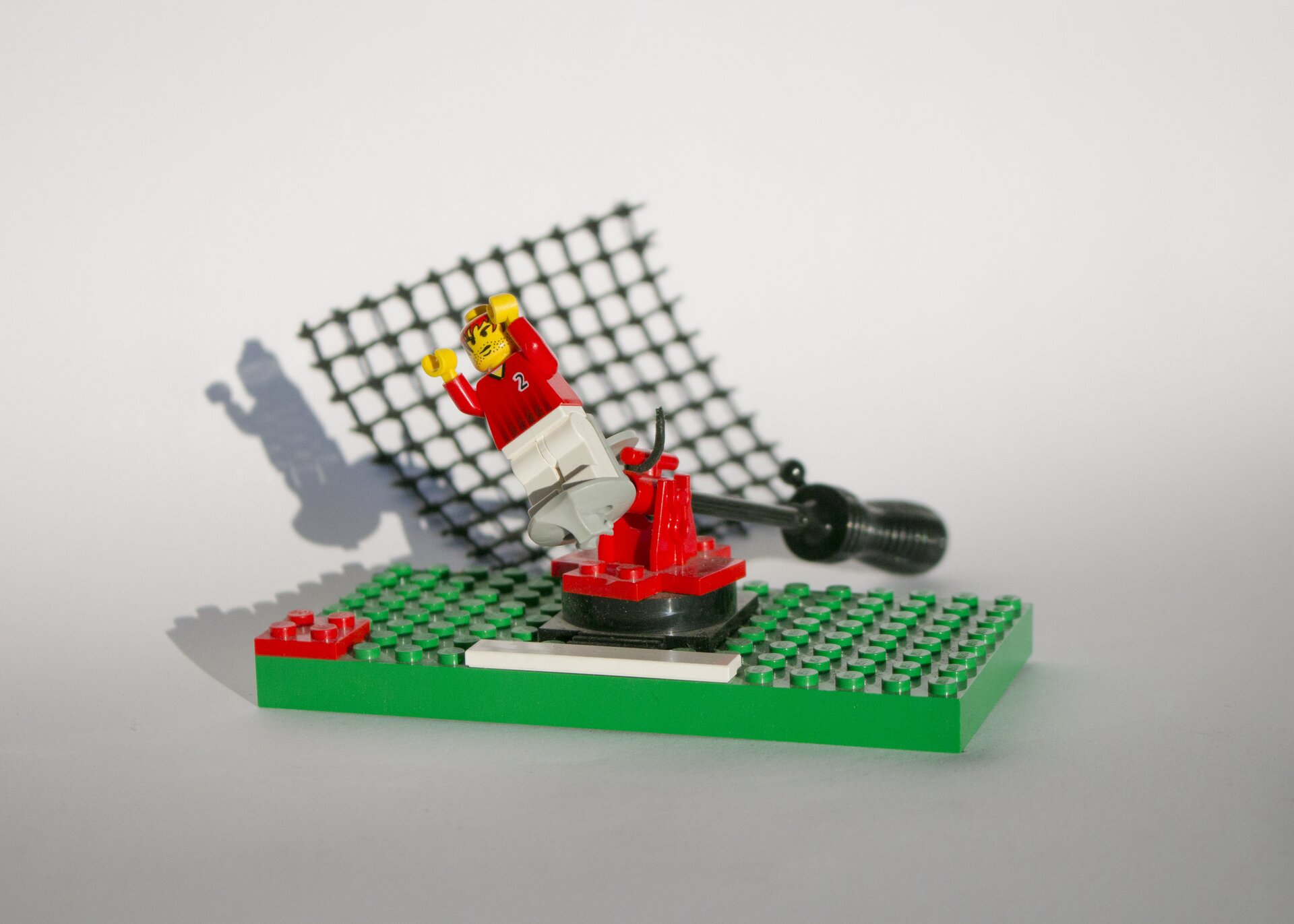 Ilustracja przedstawia fotografię „Lego”. W centrum zdjęcia znajduje się duża, zielona platforma, do której przymocowane są klocki oraz biało‑czerwono‑żółty ludzik. W tle znajduje się czarny śrubokręt i czarna, plastikowa siatka. Całość umieszczona jest na biało‑szarym tle.