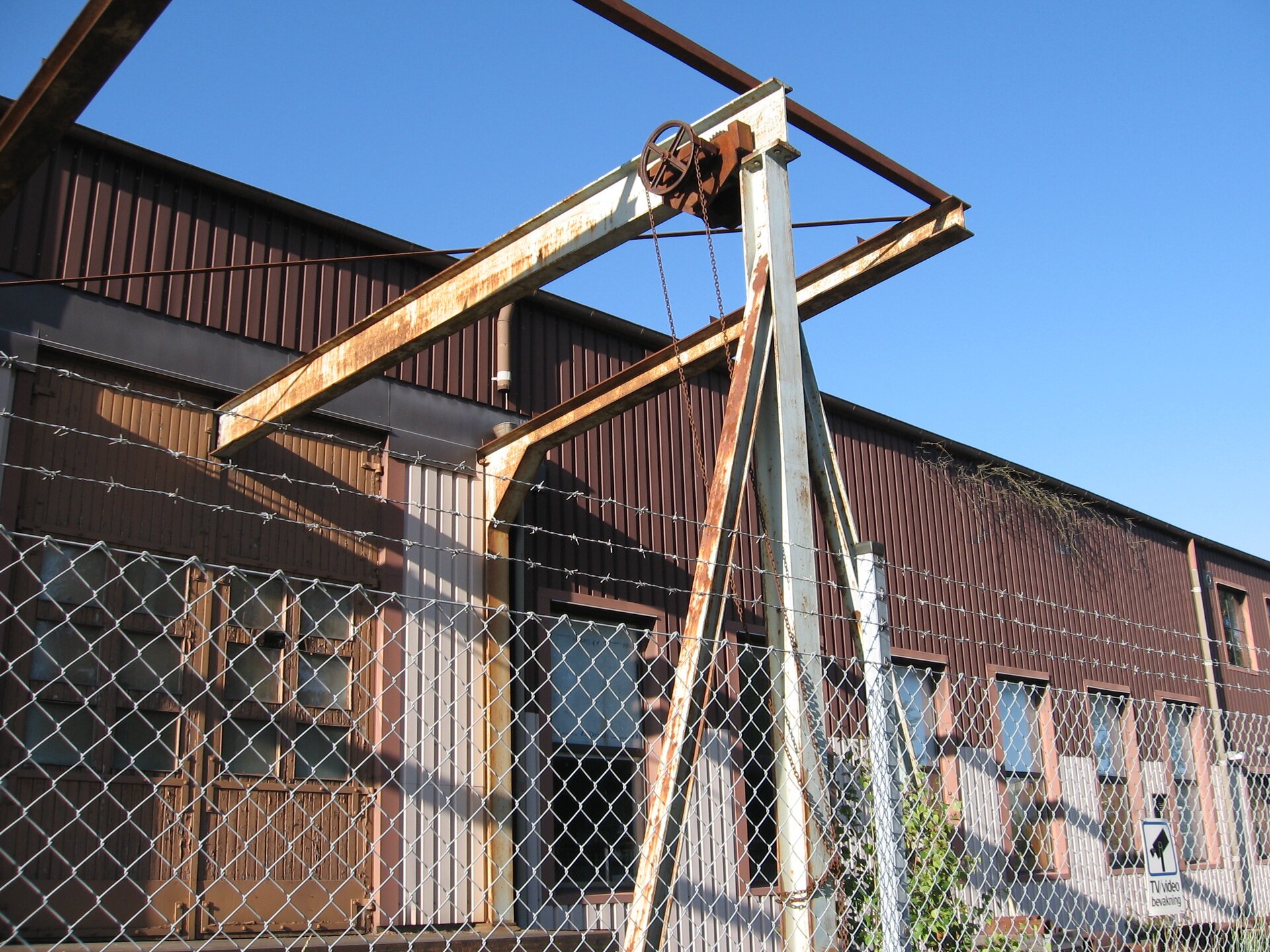 Na zdjęciu widoczna ściana budynku z przytwierdzoną stalową konstrukcją . Składa się z ramy , prowadnicy i przymocowanego do niej ruchomego koła na którym wisi łańcuch do podnoszenia ładunku