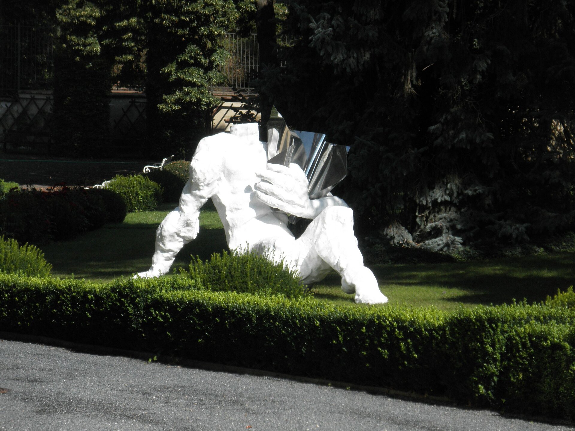 Zdjęcie przedstawia dzieło autorstwa Tomasza Koclęgi, pod tytułem: Rzeźba figuratywna. Na fotografii znajduje się biała rzeźba, przedstawiająca postać bez głowy. W jej lewej ręce znajduje się srebrny przedmiot, przypominający zwinięty arkusz. Postać siedzi na trawie z ręką opartą o ziemię. Rzeźba umieszczona jest w plenerze, gdzie rosną zielone krzewy oraz drzewa.