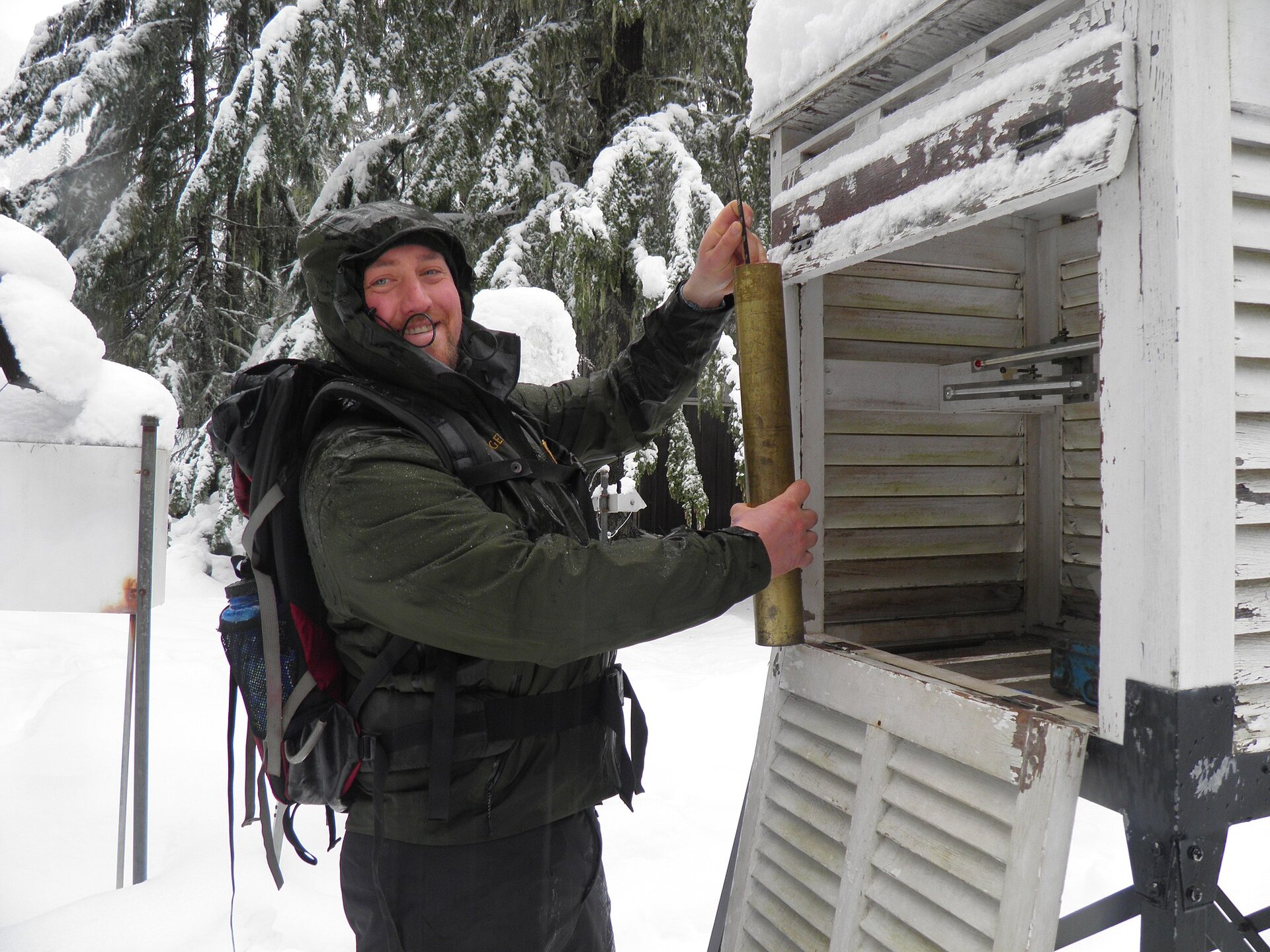 Zdjęcie przedstawia mężczyznę przy klatce meteorologicznej. Klatka jest otwarta. Mężczyzna coś w niej montuje. Wokół widać śnieżny krajobraz. Mężczyzna jest ubrany ciepło, w zimową kurtkę.
