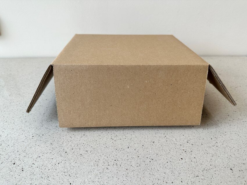 Zdjęcie przedstawia małe pudło szare z tektury falistej, o lekko rozłożonych bokach.