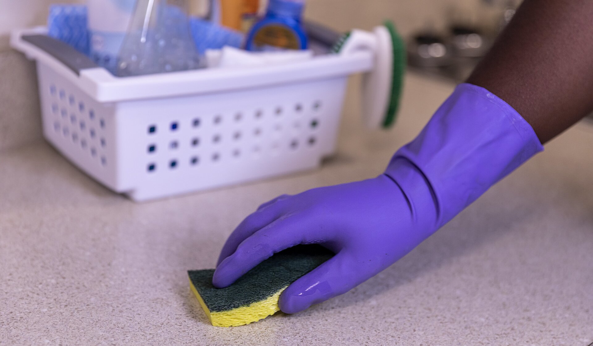 Na pierwszym planie fotografii widać rękę odzianą w gumową rękawicę ochronną. Osoba ta trzyma gąbkę do czyszczenia, którą wyciera blat. W tle widać koszyk ze środkami czystości.