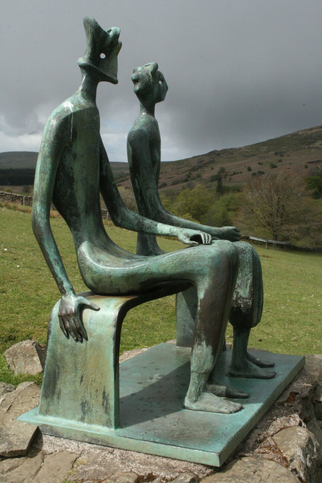 Zdjęcie przedstawia rzeźbę w kolorze zielono‑brązowym. Przedstawia dwie postacie, mężczyznę i kobietę, siedzących obok siebie na ławce. Zwrócone są lekko w lewo. Po lewej stronie znajduje się postać mężczyzny i jest wyższa. Ma szerokie ramiona i wąską talię oraz biodra. Cała sylwetka jest spłaszczona. Lewą rękę ma na kolanach, a prawą na brzegu ławki. Jego bose stopy spoczywają na ziemi. Po prawej stronie znajduje się postać kobiety. Jej sylwetka również jest spłaszczona, ale są zaznaczone wypukłe piersi. Prawą dłoń trzyma na kolanach, a bose stopy na ziemi. Podstawa rzeźby ma kształt prostokąta. Jest wykonana z takiego samego materiału jak rzeźba. Dookoła są szare kamienie i zielona trawa. W tle są pagórki porośnięte roślinnością i zachmurzone niebo. 