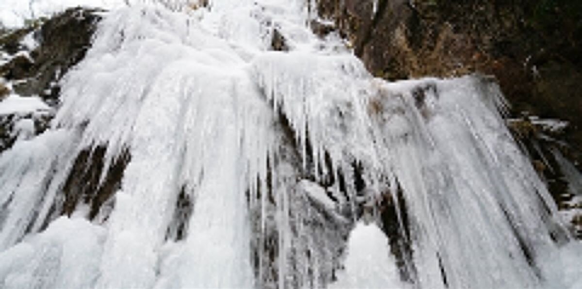 Ilustracja przedstawia fotografię skały oraz wodospad. Spływająca z niego woda jest zamarznięta, powstały długie, ostre sople o różnej grubości. Zdjęcie zostało zrobione w ciągu dnia, podczas zimy.  