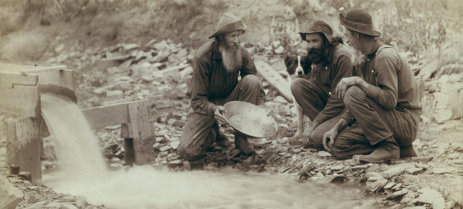 Zdjęcie przedstawia trzech brodatych mężczyzn, którzy kucają przy strumieniu wypływającym z drewnianej rynny. Postacie są ubrane w stroje robocze, mają na głowach kapelusze. Pierwszy z lewej trzyma w dłoniach metalową, płaską misę. Obok siedzi pies. Za nimi widać kamieniste podłoże oraz trawy porastające zbocze.  