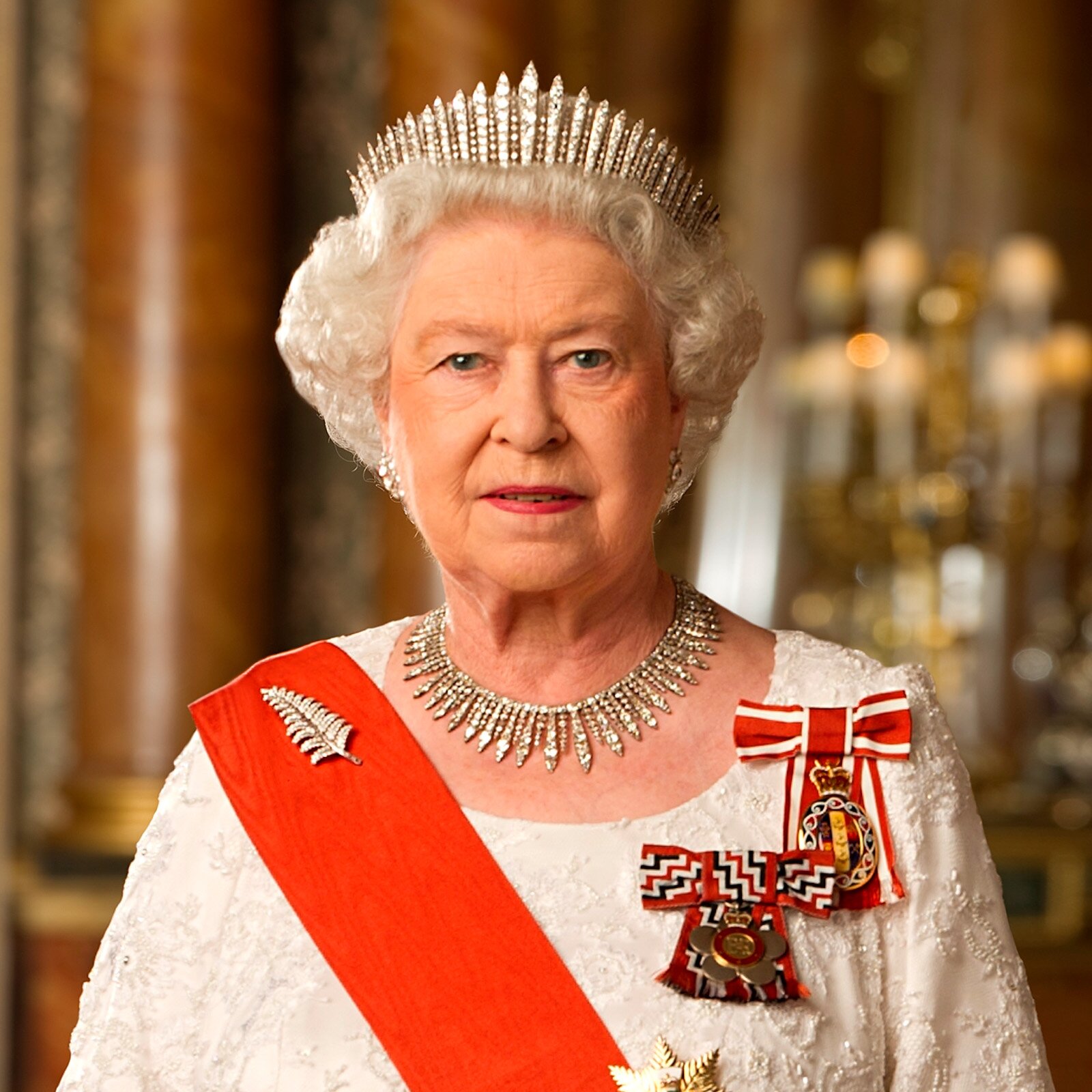 Zdjęcie przedstawiające kobietę o siwych włosach ubraną w elegancki strój. Na głowie ma koronę, a przez ciało przewieszoną czerwoną szarfę. Na piersi przywieszone ma odznaki.