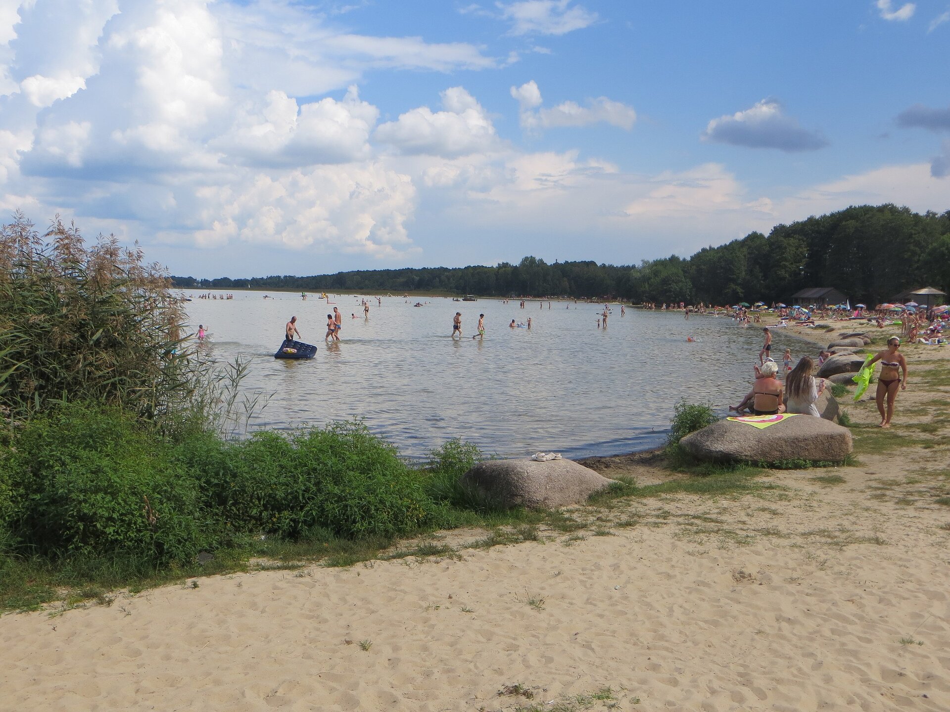 Na pierwszym planie znajduje się piaszczysta plaża, na której wypoczywają ludzie. W tle okalający jezioro las. 