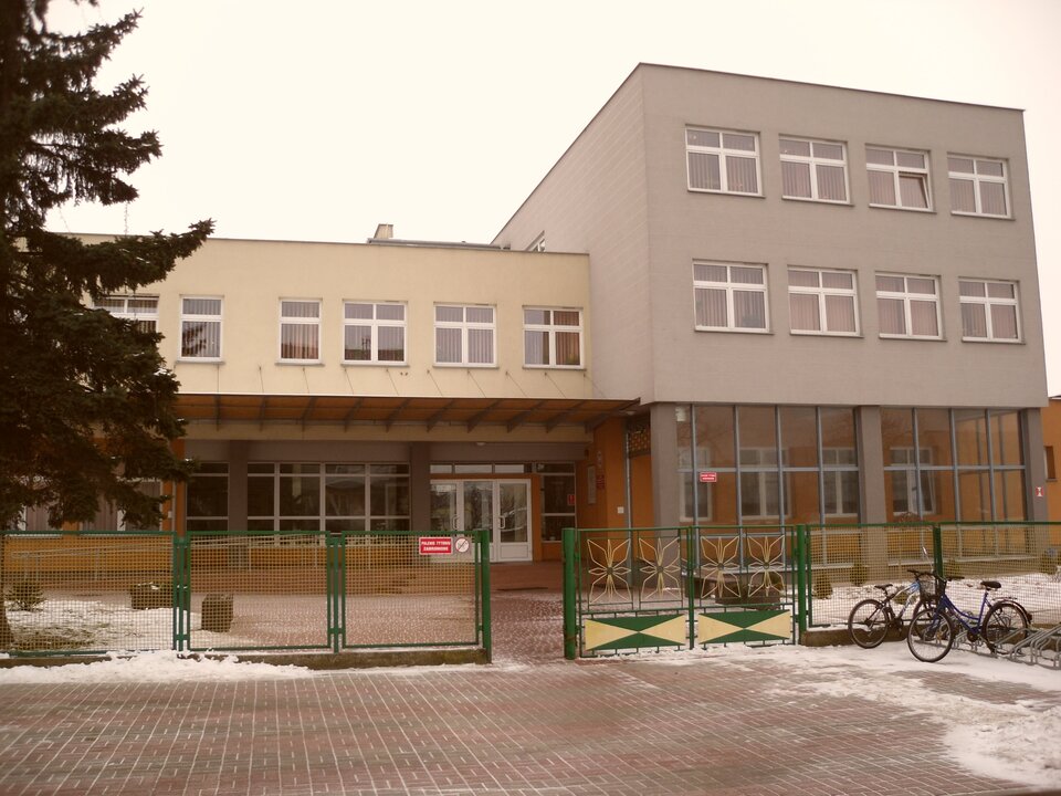 Ilustracja przedstawia budynek trzykondygnacyjny w kolorze szarym. Drzwi i okna w kolorze białym. Plac przed budynkiem wyłożony jest kostką brukową. Budynek ogrodzony jest metalowym płotem wykonanym z siatki w kolorze zielonym.