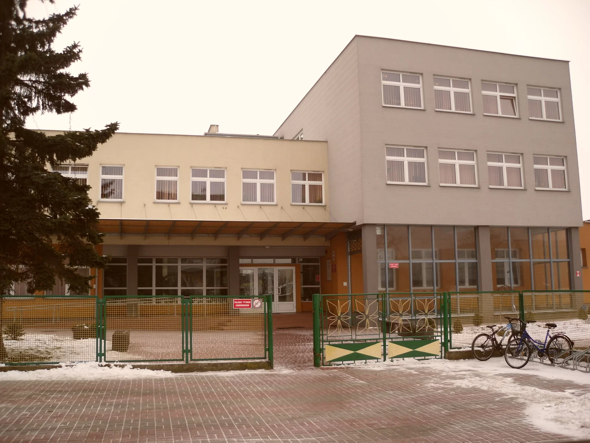 Ilustracja przedstawia budynek trzykondygnacyjny w kolorze szarym. Drzwi i okna w kolorze białym. Plac przed budynkiem wyłożony jest kostką brukową. Budynek ogrodzony jest metalowym płotem wykonanym z siatki w kolorze zielonym.