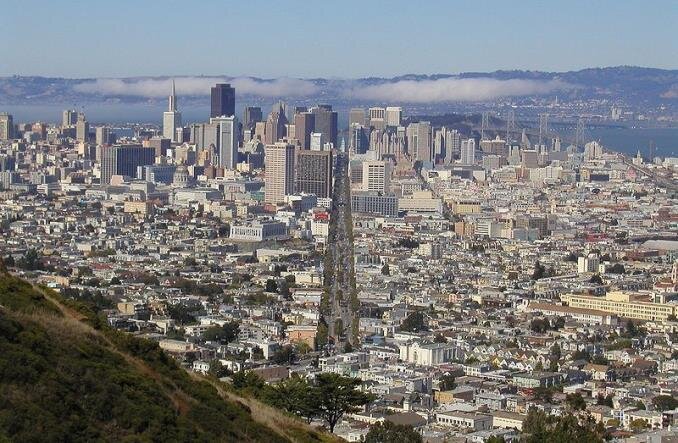 Zdjęcie przedstawia widok ze wzgórza na metropolię. Miasto ma bardzo gęstą zabudowę. Na pierwszym planie są niskie budynki, w oddali wieżowce. Pomiędzy budynkami znajduje się szeroka aleja. W tle jest zatoka i wzniesienia.