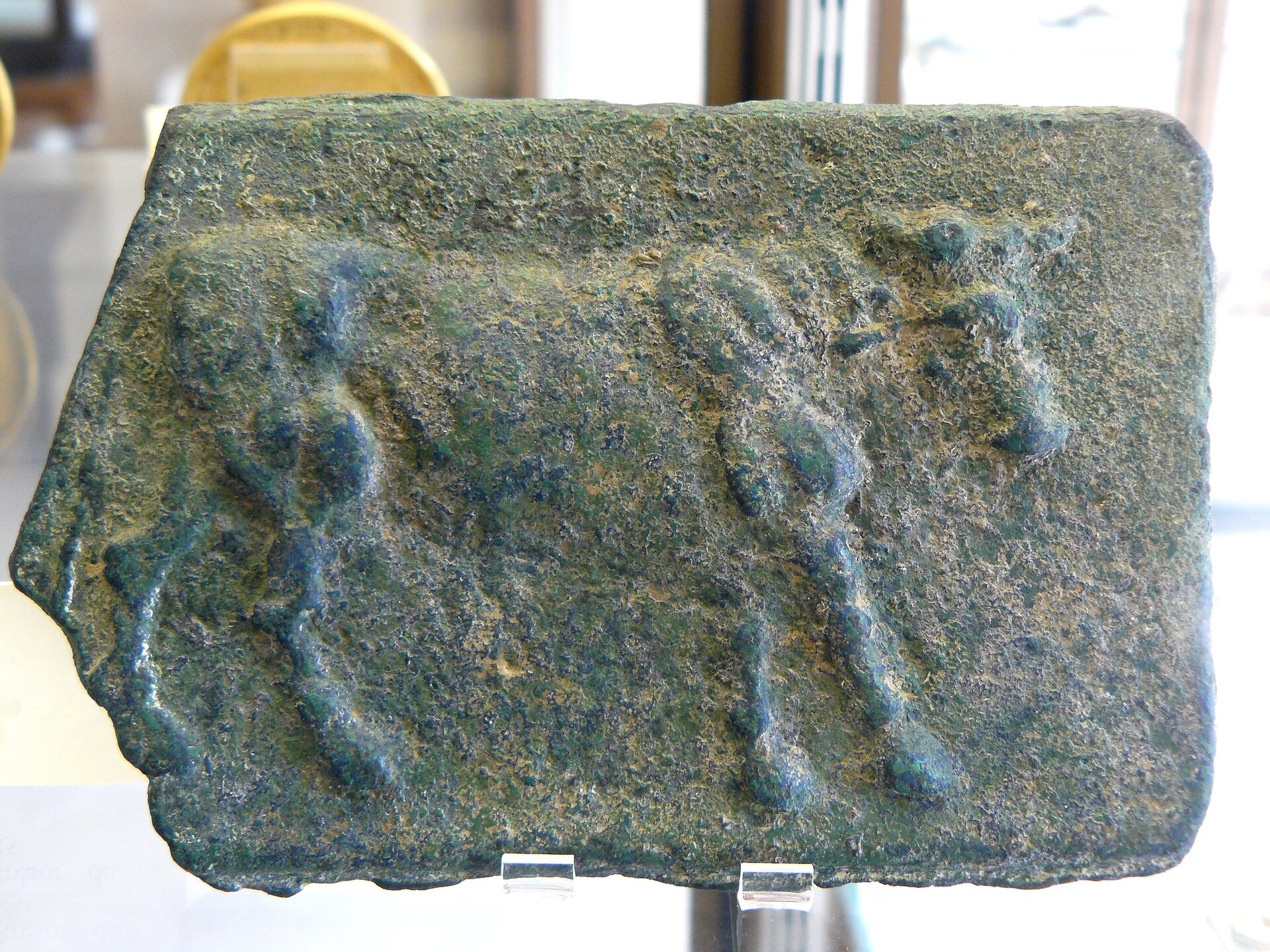 Ilustracja przedstawia płytkę z brązu, na której widnieje wizerunek krowy w formie płaskorzeźby. Całość jest koloru ciemnoszarego, zielonkawego, z brązowymi przebarwieniami. Zwierzę przedstawiono z boku. Skierowane jest ku prawej stronie.