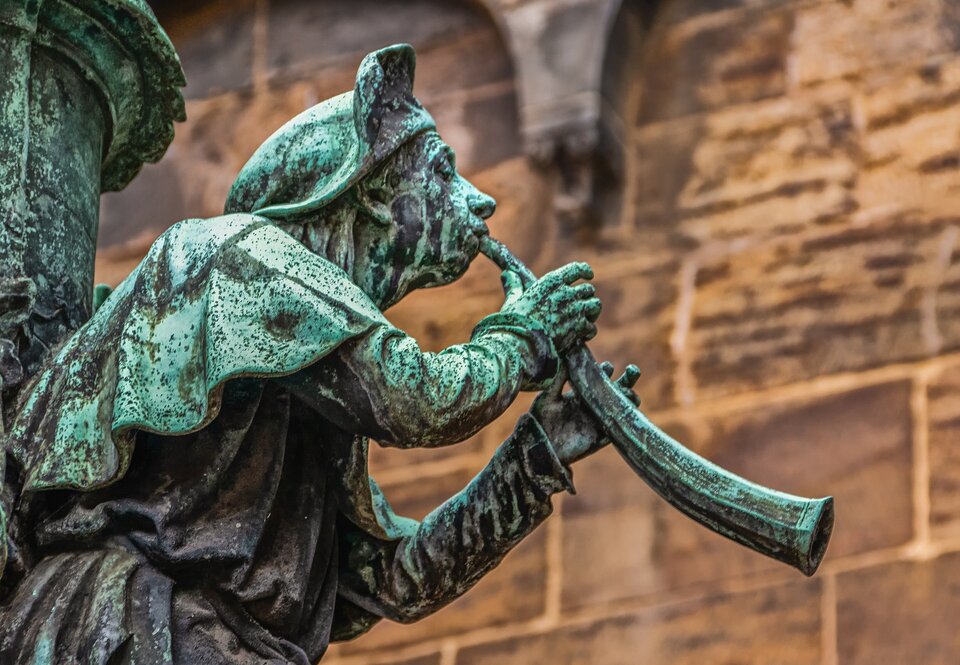 Zdjęcie przedstawia fragment pomnika. Ukazany jest średniowieczny muzyk grający na rogu.