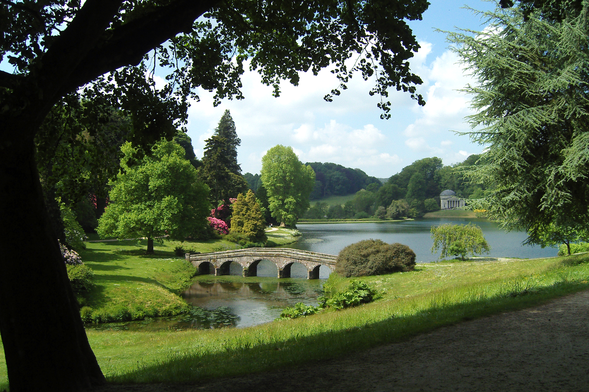 Fotografia prezentuje park położony nad wodą z różnymi drzewami oraz krzewami.