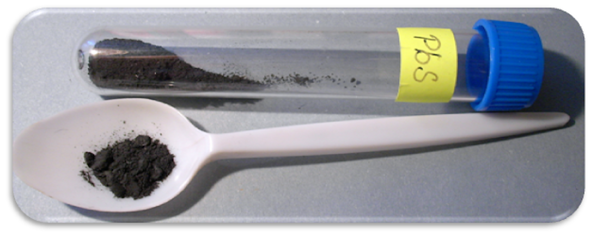 Zdjęcie przedstawiające probówkę z niebieską nakrętką, w której znajduje się czarna, drobnokrystaliczna substancja - siarczek ołowiu. Probówka jest opisana PbS. Obok probówki leży plastikowa łyżeczka, na której jest pewna ilość siarczku ołowiu. 