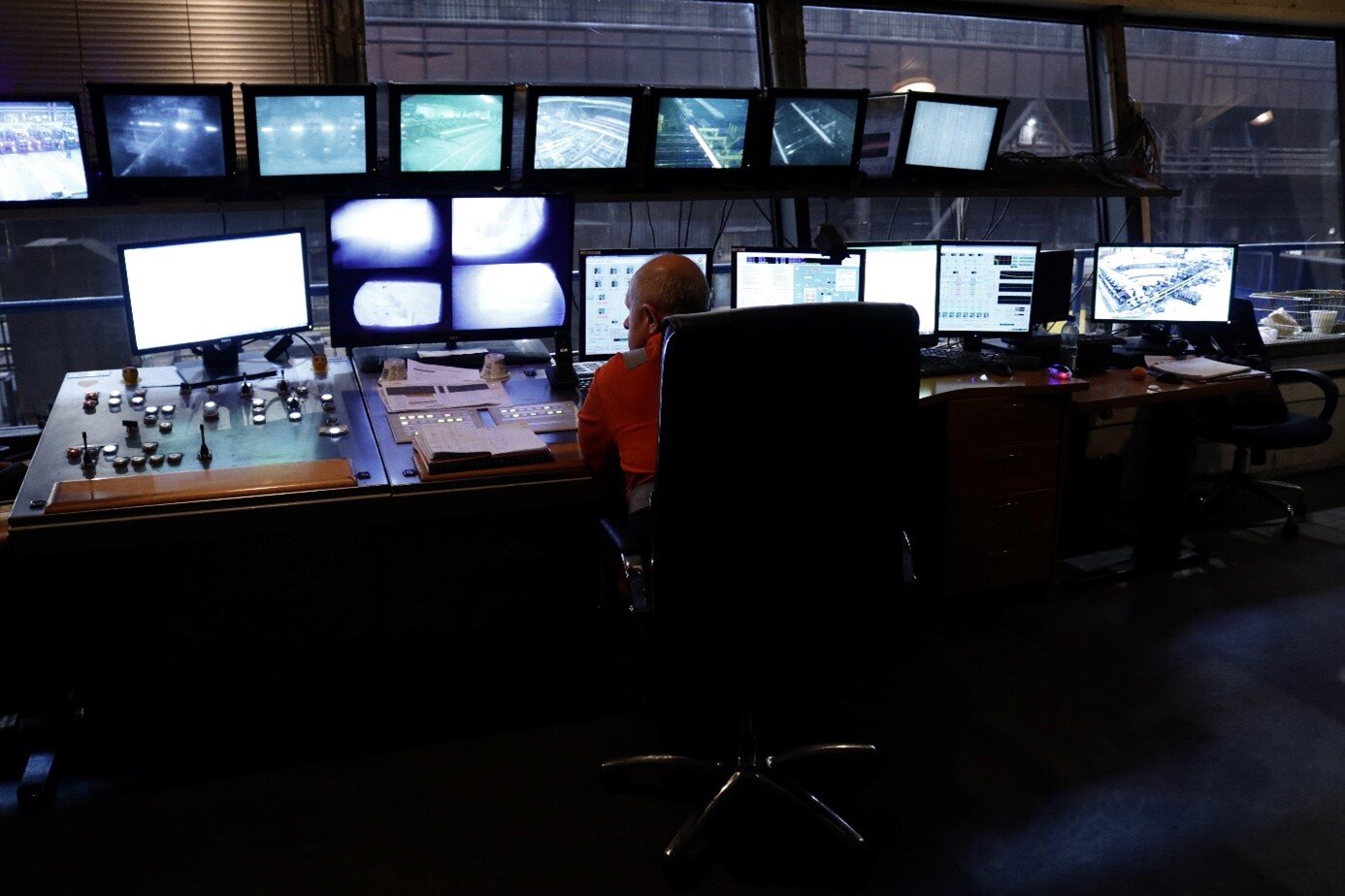 Zdjęcie przedstawia pulpit sterowniczy w kabinie operatorskiej linii walcowniczej. Na pierwszym planie zdjęcia znajduje się starszy mężczyzna siedzący na fotelu przy pulpicie sterowniczym. Mężczyzna ubrany jest w pomarańczowy strój pracowniczy. Przy biurku, na którym znajduje się pulpit sterowniczy znajduje się wiele monitorów, część z nich podwieszona jest przy oknie. Część monitorów pokazuje obraz  z kamer, a pozostałe przedstawiają parametry urządzeń. Na drugim planie widoczne są okna, a za nimi hala linii walcowniczej.