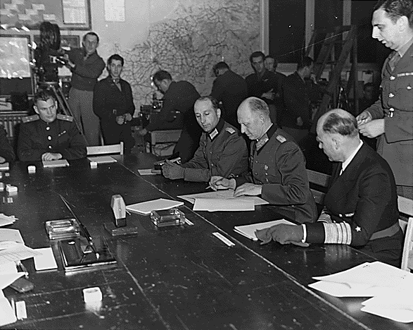 Na fotografii widać pomieszczenie, w którym przy dużym stole zasiadają niemieccy, radziecki i amerykańscy oficerowie. Podpisują dokumenty. Za ich plecami widać operatora kamery, który filmuje zdarzenie. W kącie sali stoją inni mężczyźni. 