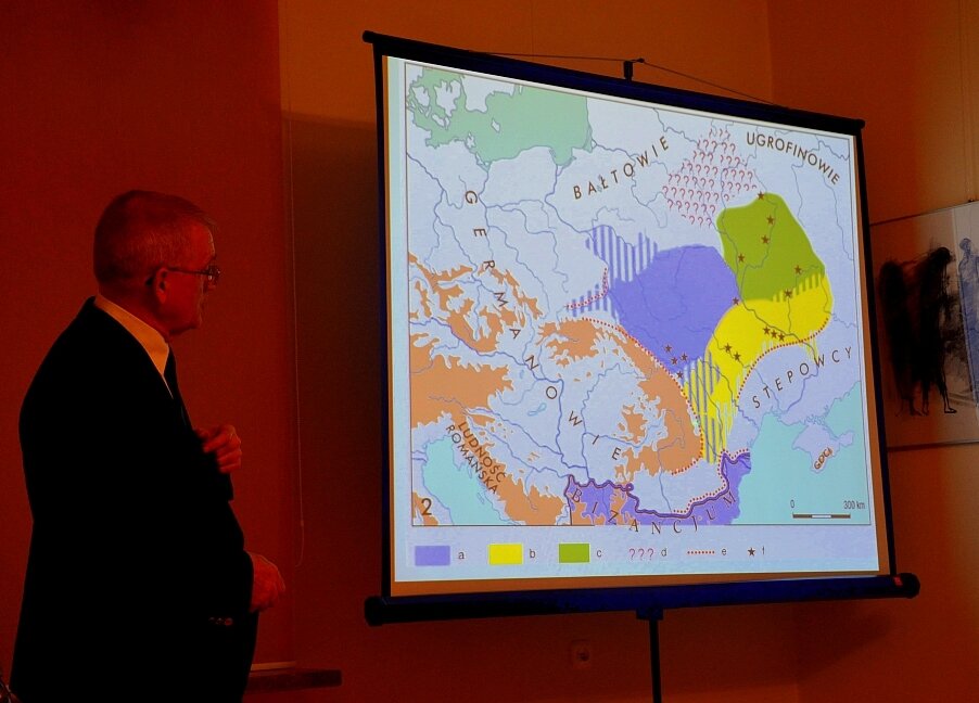 Na zdjęciu znajduje się mężczyzna w marynarce prowadzący wykład. Spogląda on na mapę, która jest wyświetlana na ekranie do projektora. Na mapie różnymi kolorami zaznaczono pewne fragmenty Europy środkowowschodniej.