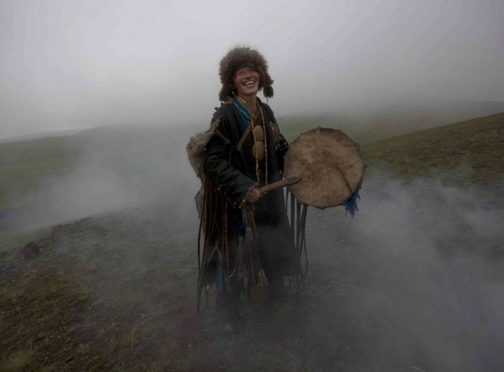 Ilustracja przedstawia szamana z Mongolii, który stoi na wzgórzu i uderza o bęben. W tle jest dużo dymu - w czasie ceremonii przebłagiwania duchów. Mężczyzna ubrany jest w strój ze skóry zwierzęcej, przy rękawach ma frędzle o długości do ziemi. W trakcie ceremonii jest bardzo uśmiechnięty, widoczne są białe zęby.