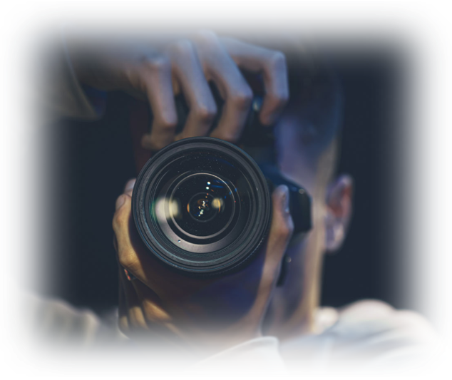 Zdjęcie przedstawia fotografującego. Kadr obejmuje zbliżenie na aparat fotograficzny, trzymany w dłoniach wraz z obiektywem skierowanym na wprost widza. Za aparatem kryje się twarz fotografującego, o krótkich włosach. 