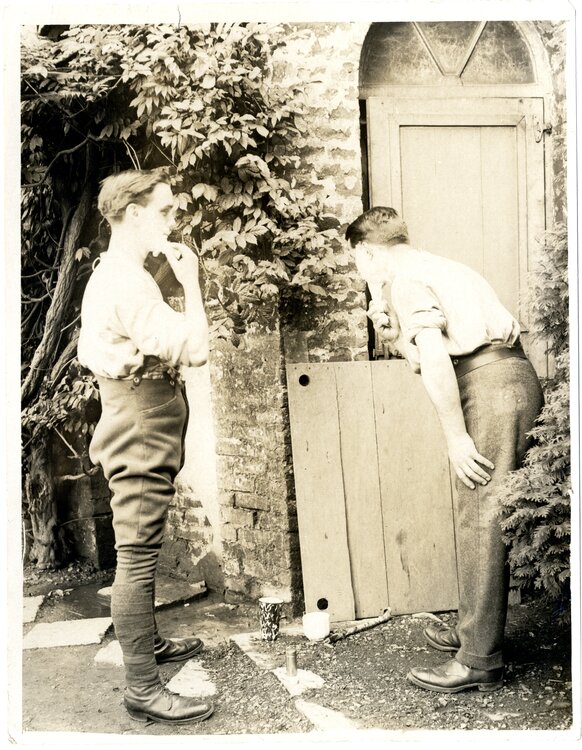 Zdjęcie przedstawia dwóch żołnierzy brytyjskich podczas golenia. Żołnierz z lewej stoi bokiem i pędzluje twarz, żołnierz z prawej tyłem, pochyla się nad deską, na której stoi malutkie lusterko. Pędzluje brodę i patrzy w lusterko. Na ziemi dwa kubki. Za mężczyznami po lewej stronie półokrągłe drzwi wejściowe, po prawej po murze pnie się milin.