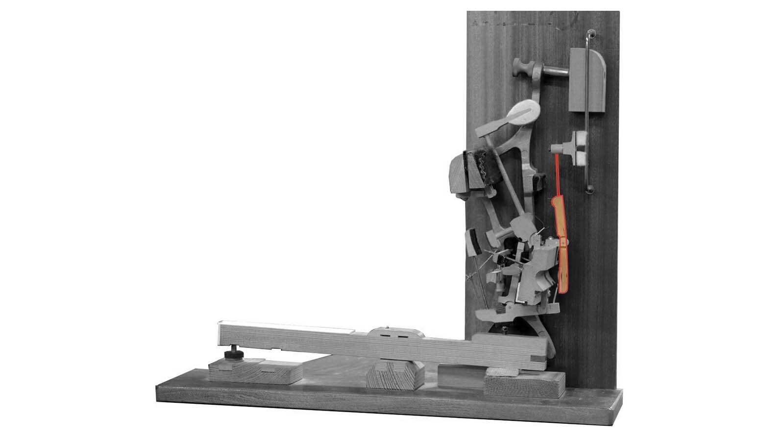 Zdjęcie przedstawia podzespół mechanizmu pianinowego lub fortepianowego jakim jest dźwignia tłumikowa. Element został zaznaczony czerwoną obwódką, a dodatkowo pozostałe elementy są w odcieniach szarości dla lepszego uwidocznienia.