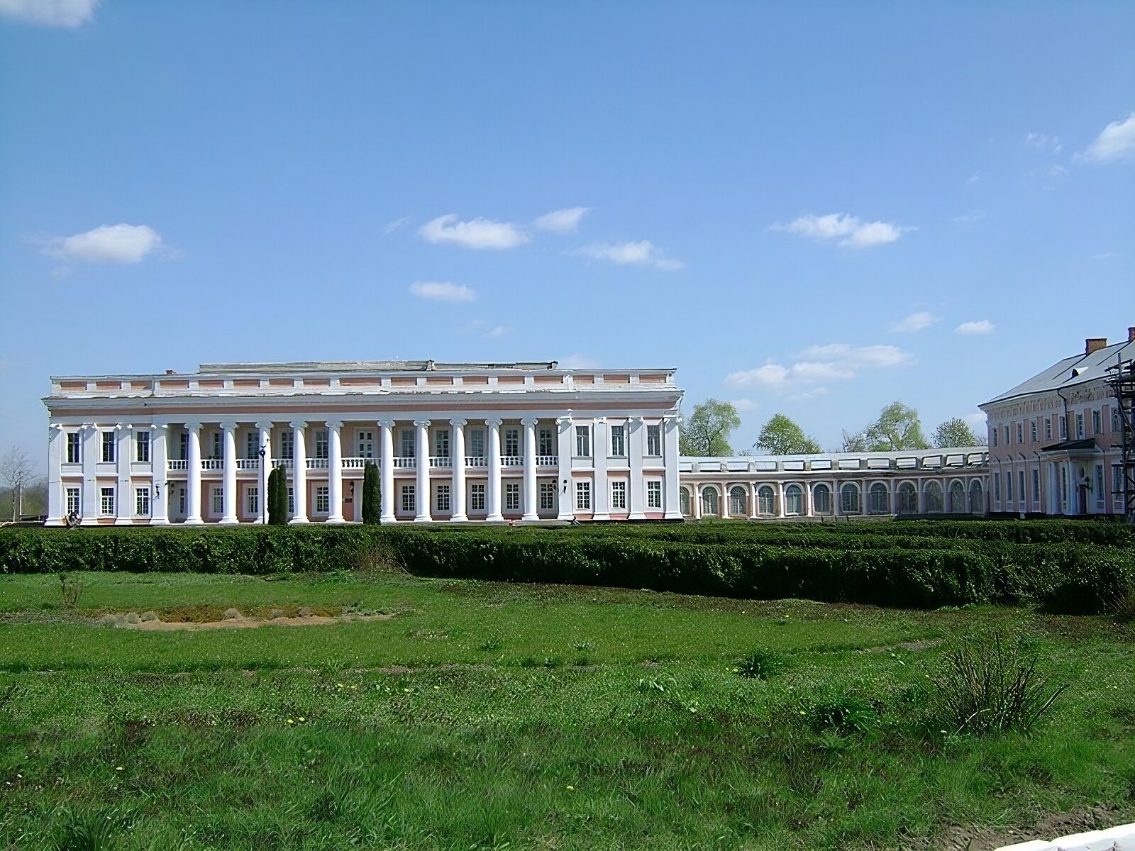 Zdjęcie przedstawia wygląd korpusu głównego pałacu w Tulczynie wraz z oszkloną, parterową galerią i fragmentem dwukondygnacyjnej, z dwukolumnowym portykiem pośrodku i dwuspadzistym dachem, oficyny z prawej strony. Pałac jest budowlą klasycystyczną dwukondygnacyjną z balkonem pośrodku na wysokości pierwszego piętra. Posiada płaski dach z fryzem ozdobnym. Fasada wejściowa pokryta jest pilastrami po bokach i kolumnami pośrodku z jońskimi kapitelami. Na pierwszym planie widoczny fragment ogrodu z trawnikiem, krzewami oraz dwoma tujami przed wejściem do głównego gmachu pałacu. 