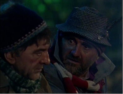 Zdjęcie przedstawia twarze dwóch mężczyzn. Po lewej stronie znajduje się prawy profil starszego mężczyzny. Jego twarz ma wyraźnie zarysowane zmarszczki. Mężczyzna ma duży nos, gęste, ciemne brwi i głęboko osadzone, opuchnięte oczy. Na głowie ma czapkę, pod szyją zawinięty szal. Po prawej stronie znajduje się mężczyzna o pociągłej twarzy. Mężczyzna ma krótki, szpakowaty zarost i wytrzeszczone oczy. Na głowie ma kapelusz, a pod nim opaskę na uszach. Ubrany jest w kurtkę, pod szyją ma zawinięty szal.