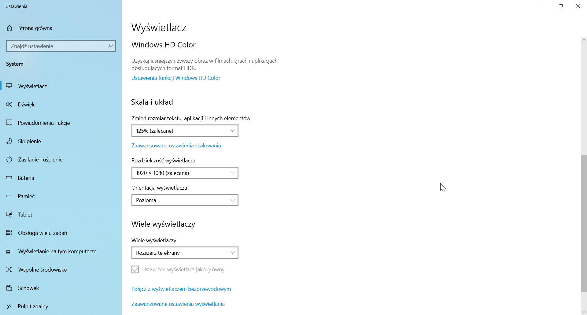 Ilustracja przedstawia okno "Ustawienia" w systemie operacyjnym Windows 10 z zakładką "System". Po lewej w panelu dostępny jest pasek wyszukiwania oraz zakładki "Strona główna", "Wyświetlacz", "Dźwięk", "Powiadomienia i akcje", "Skupienie, "Zasilanie i uśpienie", "Bateria", "Pamięć", "Tablet", "Obsługa wielu zadań", "Wyświetlanie na tym komputerze", "Wspólne środowisko", "Schowek", "Pulpit zdalny". Otworzono zakładkę "Wyświetlacz" z widoczną dalszą częścią przewiniętą w dół. Znajdują się tutaj następujące opcje: "Windows HD Color - Uzyskaj jaśniejszy i żywszy obraz w filmach, grach i aplikacjach obsługujących format HDR" i odnośnik "Ustawienia funkcji Windows HD Color", "Skala i Układ" ze zmienianiem rozmiaru tekstu, aplikacji i innych elementów w liście rozwijalnej (ustawiona na zalecane 125%) oraz odnośnikiem do zaawansowanych ustawień skalowania, "Rozdzielczość wyświetlacza" z listą rozwijalną (ustawiona na zalecane 1920 na 1080), "Orientację wyświetlacza" z listą rozwijalną (ustawiona na poziomą) oraz "Wiele wyświetlaczy" z listą rozwijalną ustawioną na rozszerzanie ekranów i zaznaczoną niedostępną opcją "Ustaw ten wyświetlacz jako główny". Poniżej znajdują się linki "Połącz z wyświetlaczem bezprzewodowym" oraz "Zaawansowane ustawienia wyświetlania". 