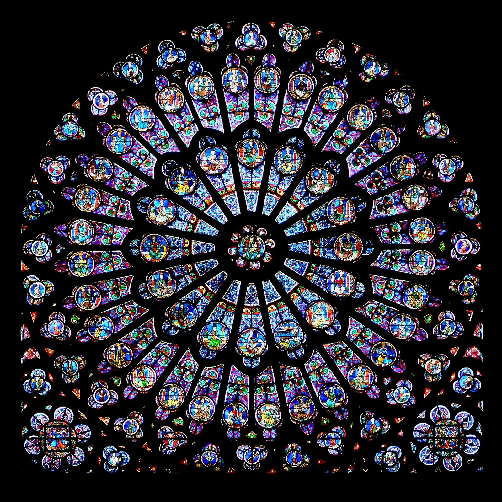 Ilustracja przedstawia zdjęcie rozety z katedry Notre Dame w Paryżu. Na zdjęciu widzimy rozbudowany witraż, Każda z części przedstawia inny motyw. Po najechaniu kursorem na ilustrację pojawi się okrąg, który przybliża fragment zdjęcia na zasadzie lupki.